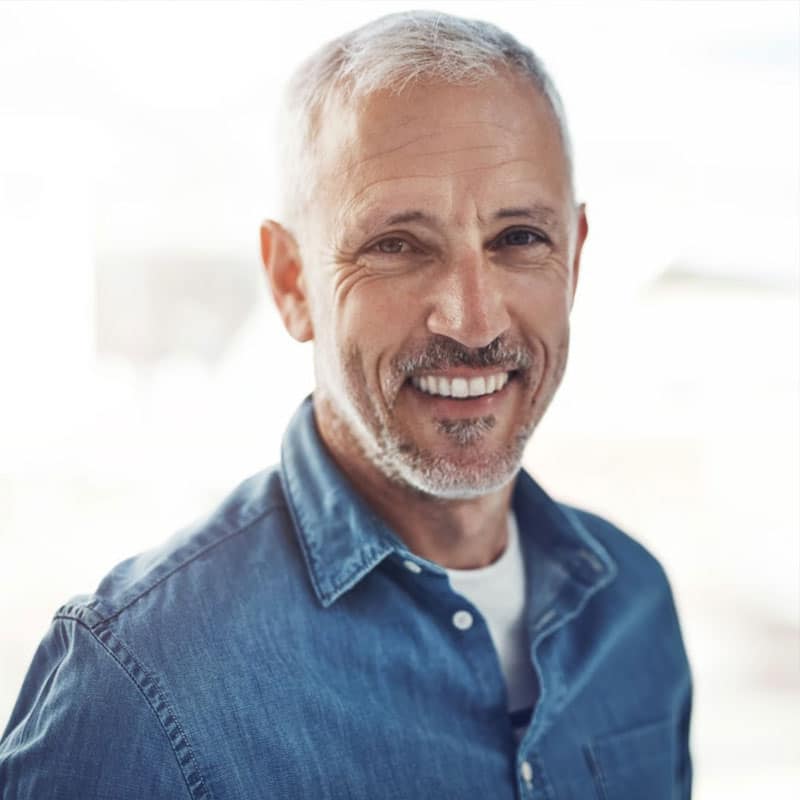 Uomo anziano con capelli grigi e sorriso amichevole, indossa camicia di jeans.